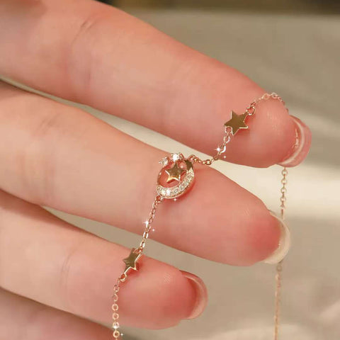 Close-up of a dainty 18k rose-gold bracelet with moon and star charms held between fingers
