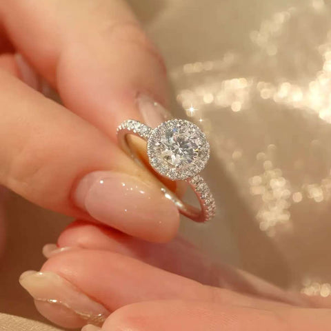 Diamond halo ring held between fingertips against shimmering fabric